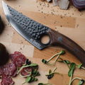 A picture of a cutting board with a Kyodai Utility Chef Knife from Seido Knives, surrounded by cured meat and leafy garnishing.