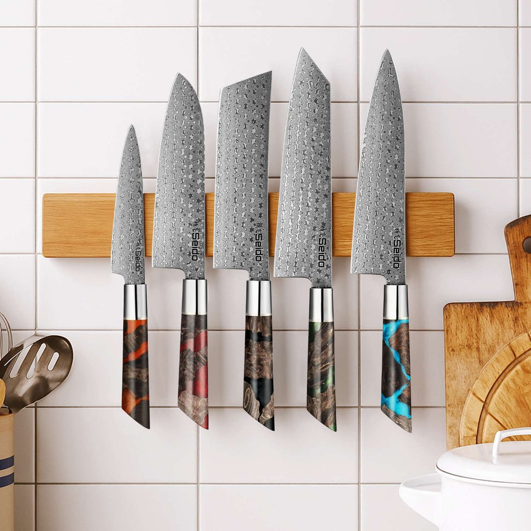 Set of five shinko series Damascus steel kitchen knives with wooden handles on a magnetic knife holder against a tiled wall.