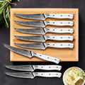 8 Takoizu serrated Damascus steak knives with white turquoise handles, arranged on a light wood cutting board against a dark background, with a small plate of butter in the corner.