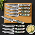 Set of 8 Awabi Damascus steel steak knives with abalone handles on a wooden block, with a small bowl of butter and herbs on a black background.