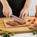 Person slicing a seared steak cleanly with an Awabi serrated steak knife on a wooden board, surrounded by vegetables and a pepper grinder.
