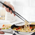 Person using long serving flat tweezers to lift noodles and vegetables from a pan at the cooktop.