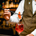 Barista garnishing a pink cocktail with a grapefruit wedge using the flat tip tweezer in a blurred bar setting.