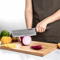 Person using a Selldo knife to slice an onion on a wooden cutting board with vegetables in the background.