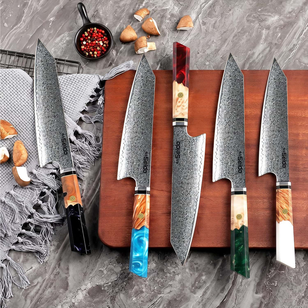Five Seido Japanese kiritsuke knives with Damascus steel blades and colorful wood-and-resin handles are arranged on a gray stone surface and a wooden board, with garlic cloves and a small bowl of peppercorns as styling props.