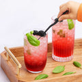 Person placing basil with offset tweezers in one of two glasses of red cocktails with ice and basil leaves on a wooden tray.