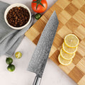 Damascus blade pattern of Shinko Kiritsuke 8.5 inch knife on a wooden cutting board with lemons, tomatoes, and coffee beans.