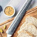 Shujin serrated edge and Damascus cladding detail with scalloped/granton reliefs, on a wooden cutting board with sliced bread.