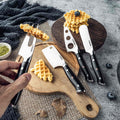 Set of cheese knives on a wooden cutting board with waffles and blueberries.