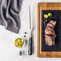 Sliced steak on a slate board with a meat fork, lime wedges, and a glass of lime juice on a marble surface.