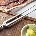 Prong detail with long tapered stainless steel tines of the meat carving fork, on a wooden surface with a bowl of lime wedges and a piece of meat in the background.