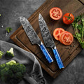 Two Damascus tengoku knives with blue handles on a wooden cutting board with sliced tomatoes and vegetables.