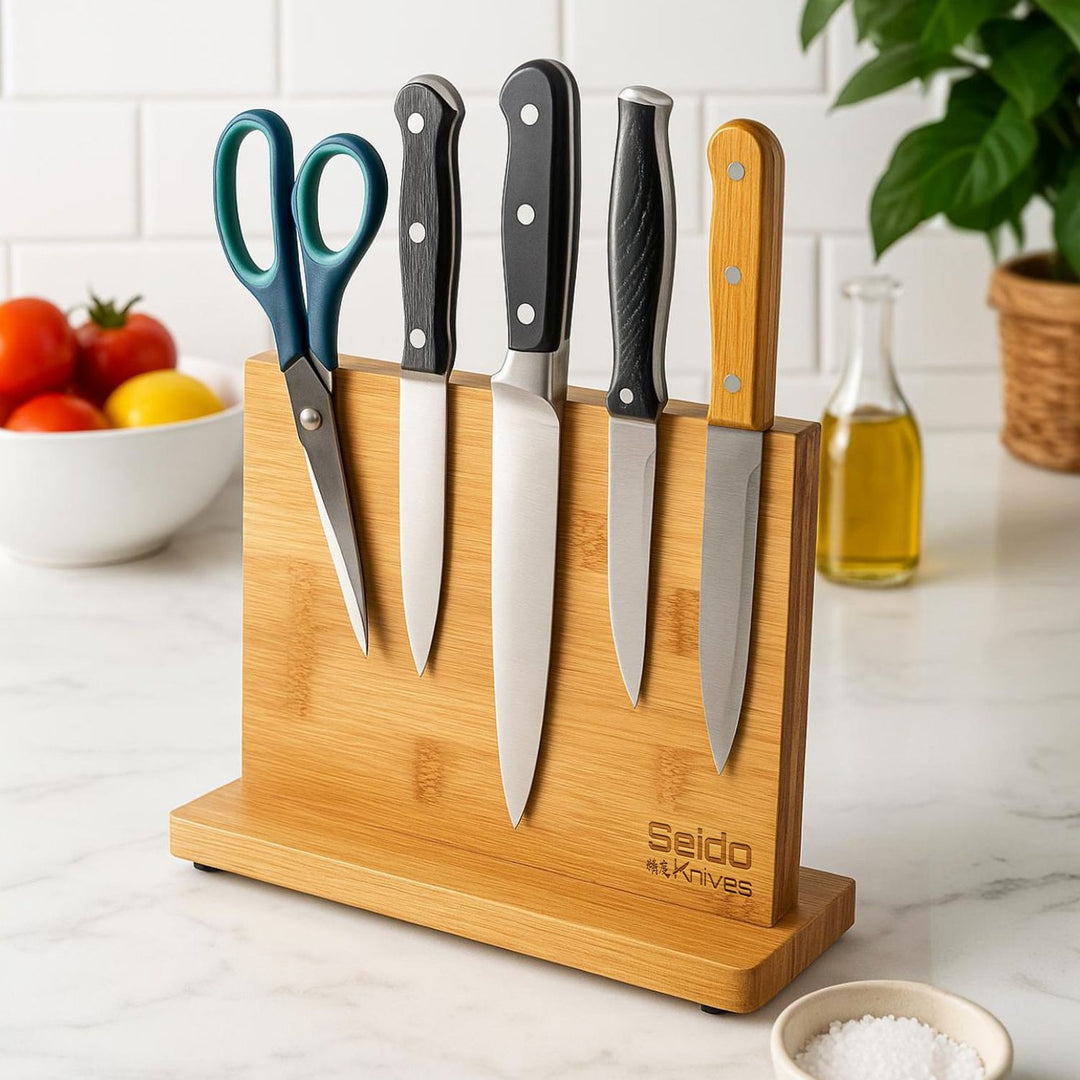 A bamboo magnetic kitchen knife block with five knives and a pair of scissors, set on a white marble countertop with a tiled backsplash, tomatoes, and a plant in the background.