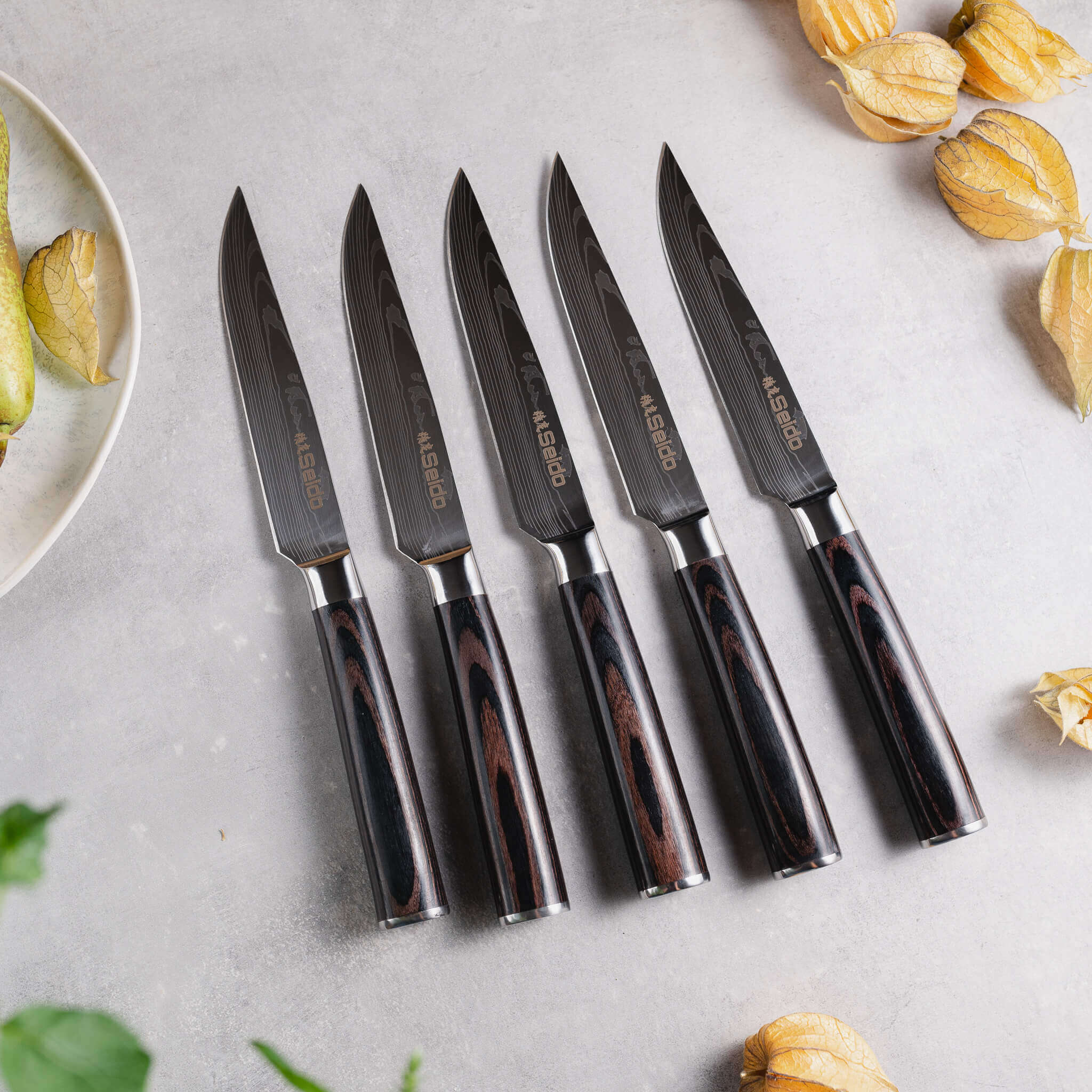 On a table, a bowl of luscious fruit is accompanied by five Seido Knives' non-serrated Straight-Edge Steak Knives.