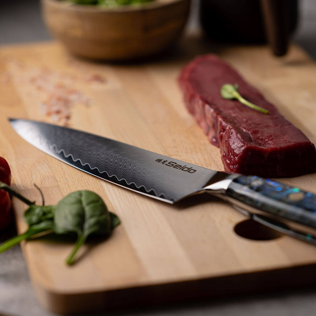 A stunning Awabi Gyuto Damascus Steel Chef Knife from Seido, resting on a cutting board with a juicy steak. 